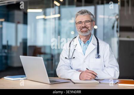 Experienced male doctor wearing a lab coat and stethoscope, smiling while sitting at his desk with a laptop and notebook, representing healthcare, wellness, and professional medical service Stock Photo