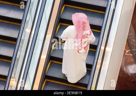 High-angle rear view of an Arab man in traditional clothing riding an escalator in a modern public building. Stock Photo