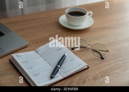 Small planner open beside coffee and glasses, pen placed diagonally on page, laptop edge visible, soft daylight creates reflective mood for weekly Stock Photo