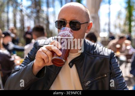 Bald man drinking beer outdoors leather jacket and sunglasses enjoying sunshine in busy beer garden with friends in background wooden table and pint Stock Photo
