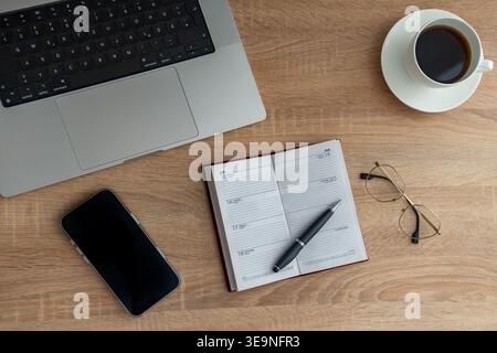 Small planner beside coffee and laptop, pen resting on open weekly spread, folded glasses and textured wood surface create calm inspired mood for idea Stock Photo