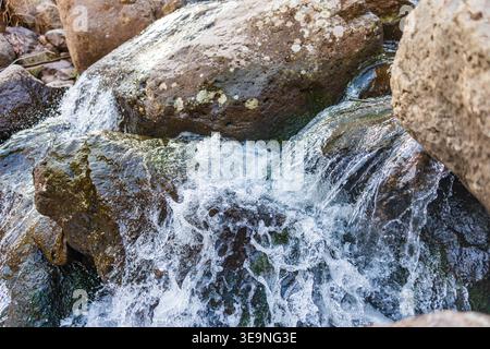 Water flows over mossy rocks in a mountain stream. Splash creates white foam against dark wet stones. Light reflects off water surface. Rocky terrain Stock Photo