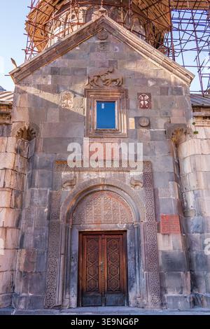 Ancient stone church with wooden door. Scaffolding surrounds upper structure. Sunlight casts shadows on textured walls. Blue window contrasts with gra Stock Photo