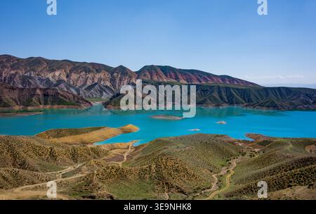 Vibrant turquoise lake surrounded by layered mountain ridges. Clear blue sky above rugged, earth-toned hills. No animals or plants visible. Aerial vie Stock Photo