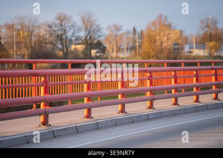 Bright red roadside safety barriers on an urban bridge with asphalt lane edge, concrete curb, and soft background trees for public works concepts Stock Photo