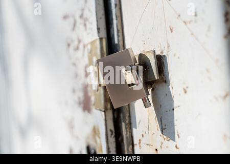 Square metal latch lock fastened on weathered white steel door with rust marks, hinge detail, and industrial security hardware close view Stock Photo