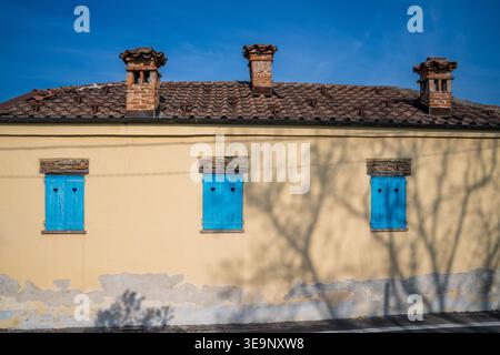 A charming yellow facade of a rural house featuring three vintage brick chimneys on a tiled roof. The windows display bright blue shutters with heart- Stock Photo