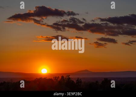 Sun setting behind distant mountains and silhouetted trees with orange sky and clouds Stock Photo