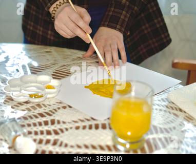 A high-quality macro shot focusing on a hand holding a brush and applying bright yellow watercolor paint to white paper. The scene captures the fluid texture of the paint and the artist's concentrated movement. The workspace includes a palette and a glass of yellow-tinted water on a lace tablecloth. Perfect for artistic backgrounds, tutorials, and creative inspiration content. mixing yellow watercolor paint on paper. Close-up of creative process and color blending. High quality photo Stock Photo