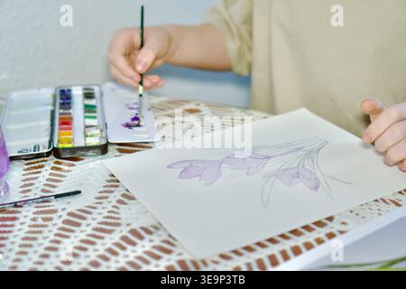 GrA high-quality medium shot focusing on an artist's hands using a brush to paint a botanical illustration of purple flowers on textured paper. In the background, a second person is visible, and the overall setup suggests a collaborative art session or workshop. A full watercolor palette and supplies are spread out on a lace tablecloth. Perfect for promoting art schools, team-building events, and creative hobby groups.up art session: people painting purple flowers with watercolors. Collaborative creative workshop and hobby education concept. High quality photo Stock Photo