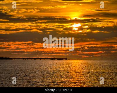 Vivid sunset with intense orange and yellow hues over the ocean, featuring a distant pier silhouette beneath dramatic layered clouds. Stock Photo