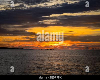 Vivid sunset with intense orange and yellow hues over the ocean, featuring a distant pier silhouette beneath dramatic layered clouds. Stock Photo