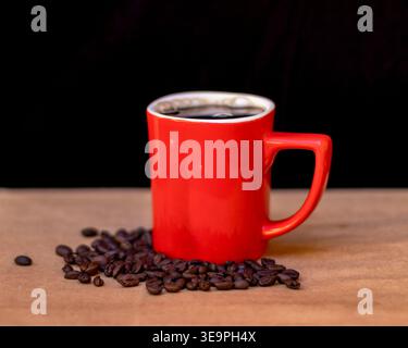 A vibrant red coffee mug filled with freshly brewed black coffee, surrounded by scattered roasted coffee beans on a wooden surface. Stock Photo