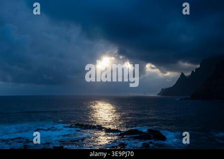 Dark clouds loom over restless ocean waves. Sunlight pierces through thick stormy sky. Rocky shore glows with gentle, scattered light. Stock Photo