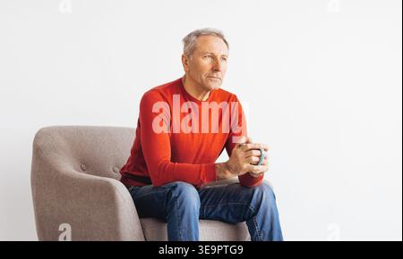Older Man Sits in Chair Holding Cup While Looking Thoughtful in Bright Room Stock Photo