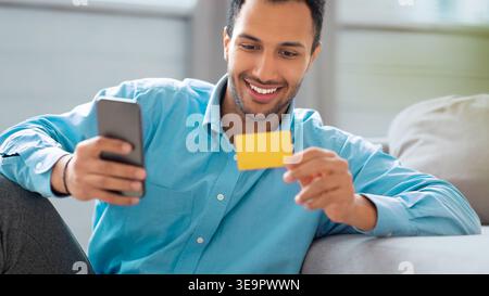 Man Sitting on a Sofa Looking at a Phone and Holding a Yellow Card While Smiling Stock Photo