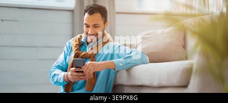 Man in Blue Shirt Sits on Sofa and Looks at Phone in Warm Indoor Setting With Natural Light Stock Photo