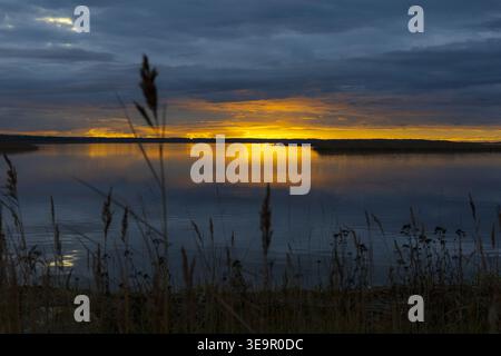 Intense sunset with glowing horizon and dark clouds over still water. Atmospheric coastal scene. Stock Photo
