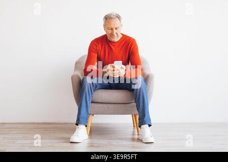 Older Man Sits on Chair and Uses Smartphone in Bright Room During Daytime Stock Photo