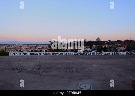 Panoramic view of Rome Skyline at sunset featuring Saint Peter's Basilica dome, with a street art banner quote in Italian on wall in foreground, Italy Stock Photo