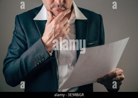 Businessman in suit thoughtfully holding a white paper sheet while resting hand on chin, reviewing documents, contracts and reports. Selective focus. Stock Photo