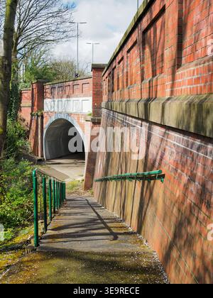 Concrete steps with green metal handrails descend beside a weathered brick retaining wall toward a sunny pedestrian tunnel archway, strong light and s Stock Photo