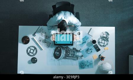 Top view of skilled engineer writing in paper while looking at tablet display green screen with turbine engine sketch design and metal gear and Stock Photo