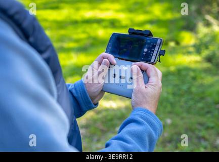 Dijon, France - April 1, 2026: A close-up of a man's fingers on a drone remote controller sticks. The phone screen shows a live video of a lonely tree Stock Photo