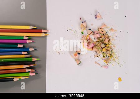 Vibrant colored pencils arranged on gray background beside white surface with fresh wood shavings and pigment remnants, showing artistic process and Stock Photo