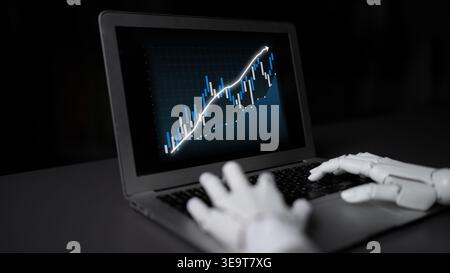 A robotic hand interacts with a laptop displaying a financial graph, illustrating the intersection of technology and finance in stock market analysis Stock Photo