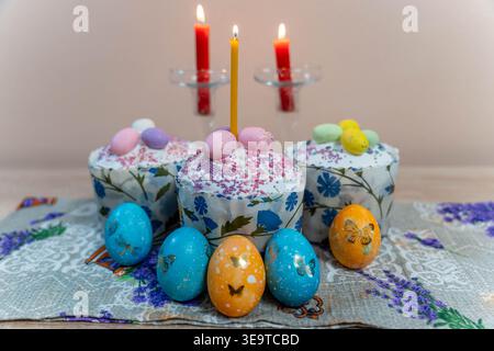 Festive Centerpiece Of Easter Cakes And Painted Eggs Arranged On Embroidered Cloth, Soft Candlelight And Floral Napkin Suggest Warm Family Gathering Stock Photo