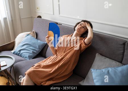 Woman lying on sofa in room fanning with paper fan suffering from head pain and summer heatstroke. Stock Photo
