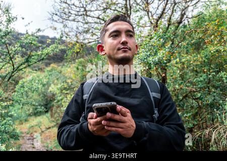 Hiker man standing on a forest trail, holding a smartphone and looking away, exploring nature and using technology Stock Photo