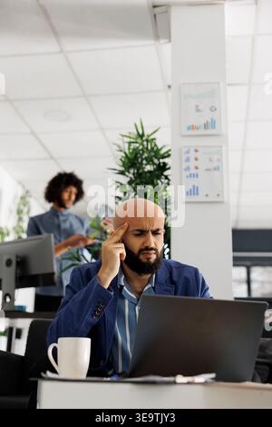 Businessman having headache while analyzing data on corporate laptop in office. Small business arab owner with professional burnout checking financial report in workplace Stock Photo