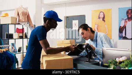 African american deliveryman asking worker to sign delivery report, discussing clients shipping detalis in shopping centre. Courier taking cardboard boxes, preparing to ship packages to customers Stock Photo