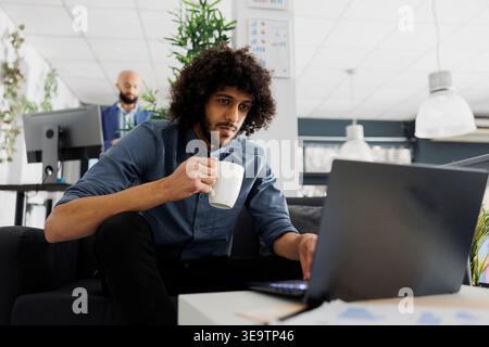 Young arab comapny employee drinking coffee and using corporate laptop in business office. Executive manager holding tea mug and solving work tasks on computer in coworking space Stock Photo