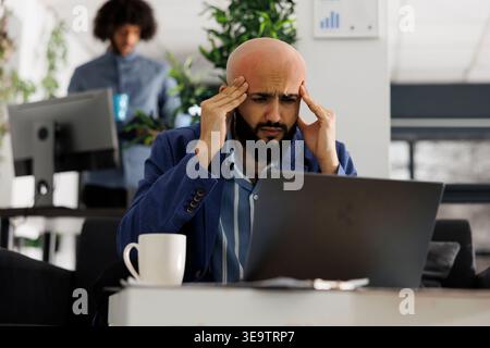 Start up product manager with headache working to meet deadlines in business office. Arab company entrepreneur suffering from migraine and rubbing temples at workplace in coworking space Stock Photo