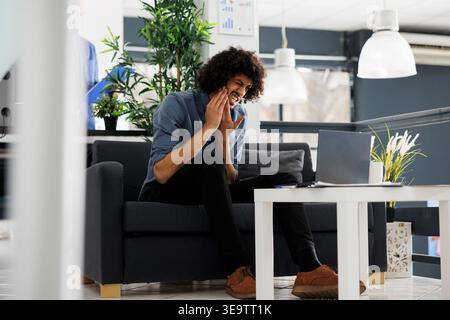 Young arab business manager suffering from toothache in start up office. Company employee having tooth pain while working at strategy on laptop in corporate coworking space Stock Photo
