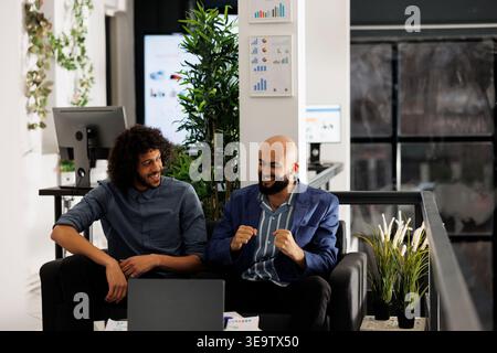Excited arab managers celebrating sales increase while checking report on laptop in business office. Smiling startup employees team happy about project success in coworking space Stock Photo