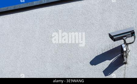 Security surveillance camera mounted on a building wall casting shadow in sunlight, minimalist security concept with copy space. Stock Photo