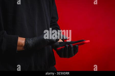 Anonymous scammer or hacker wearing black gloves using smartphone on red background. Online fraud, cybercrime, mobile hacking, identity theft and phis Stock Photo