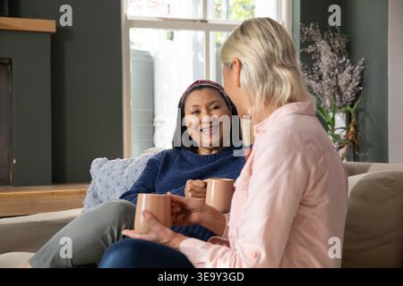 Diverse female friends sitting on beige sofa holding matching ceramic mugs near window and pillow Stock Photo