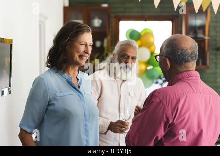 Diverse friends chatting at home, senior Indian man holding wine glass near balloons and cabinets Stock Photo