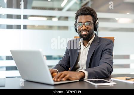 Professional man wearing glasses and headphones, smiling while typing on a laptop at an office desk, symbolizing productive remote work and modern communication technology Stock Photo