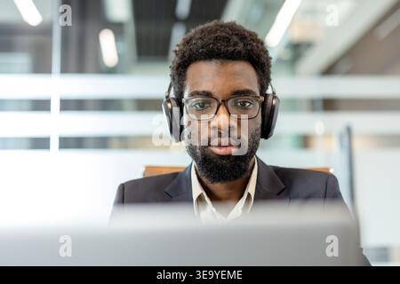 Young businessman wearing headphones and glasses is focused on his laptop during a virtual meeting or remote work session in a modern office, embodying concentration and productivity Stock Photo