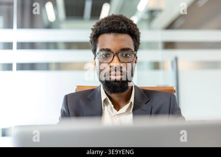 Confident male executive in glasses and a suit seated at a modern office desk, looking directly at camera with a serious, focused expression conveying leadership and determination Stock Photo