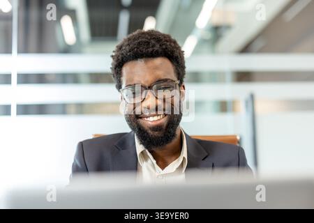 Confident business person with eyeglasses and a beard smiling while looking down at a laptop screen in a bright, contemporary workspace, representing success and productivity Stock Photo
