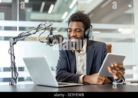 Smiling podcaster wearing headphones and recording audio content, using a microphone, laptop, and digital tablet for an interview or broadcast session in a contemporary office setting Stock Photo