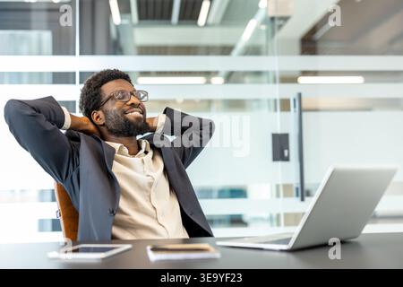 Satisfied businessman leaning back in his chair with hands behind head, smiling in a modern office, feeling accomplished after completing tasks on his laptop Stock Photo