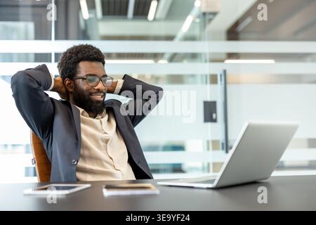 Young professional man with a beard and eyeglasses leaning back in his chair, smiling and looking at a laptop while taking a break from work in a bright, contemporary office environment Stock Photo